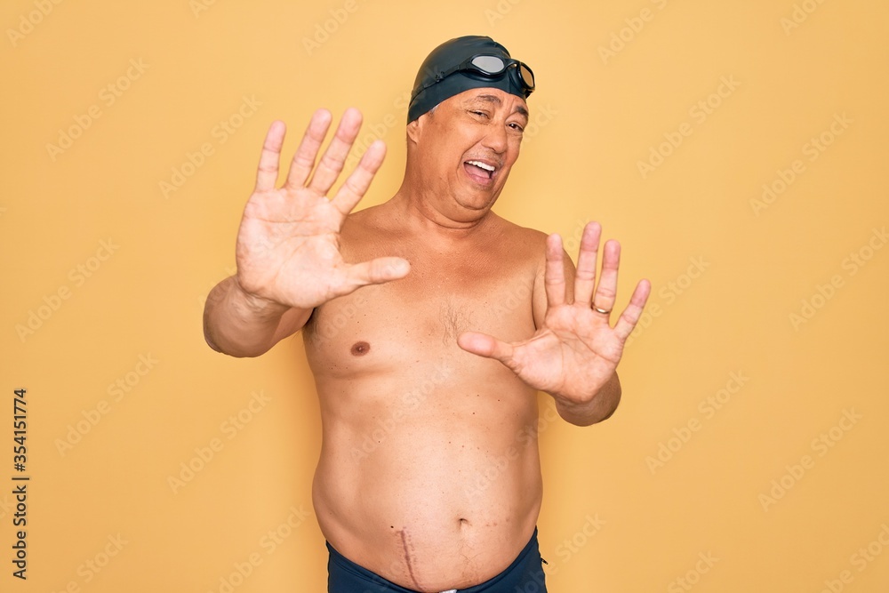 Middle age senior grey-haired swimmer man wearing swimsuit, cap and goggles afraid and terrified with fear expression stop gesture with hands, shouting in shock. Panic concept.
