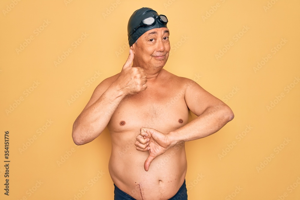 Middle age senior grey-haired swimmer man wearing swimsuit, cap and goggles Doing thumbs up and down, disagreement and agreement expression. Crazy conflict