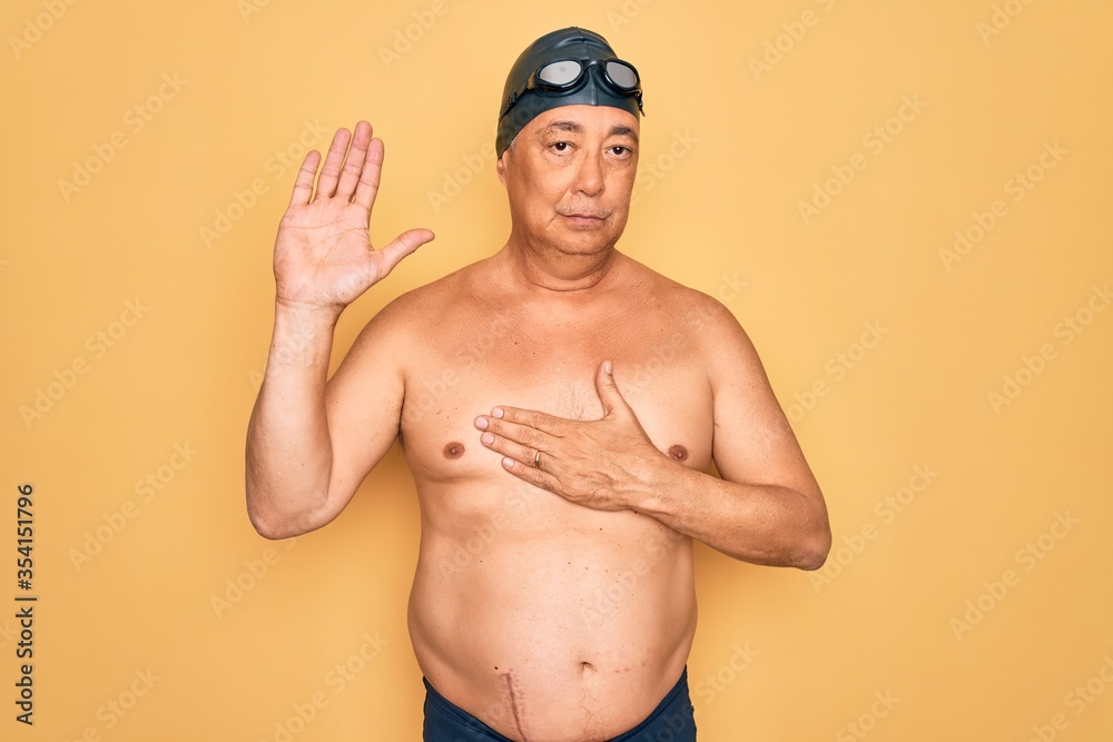 Middle age senior grey-haired swimmer man wearing swimsuit, cap and goggles Swearing with hand on chest and open palm, making a loyalty promise oath