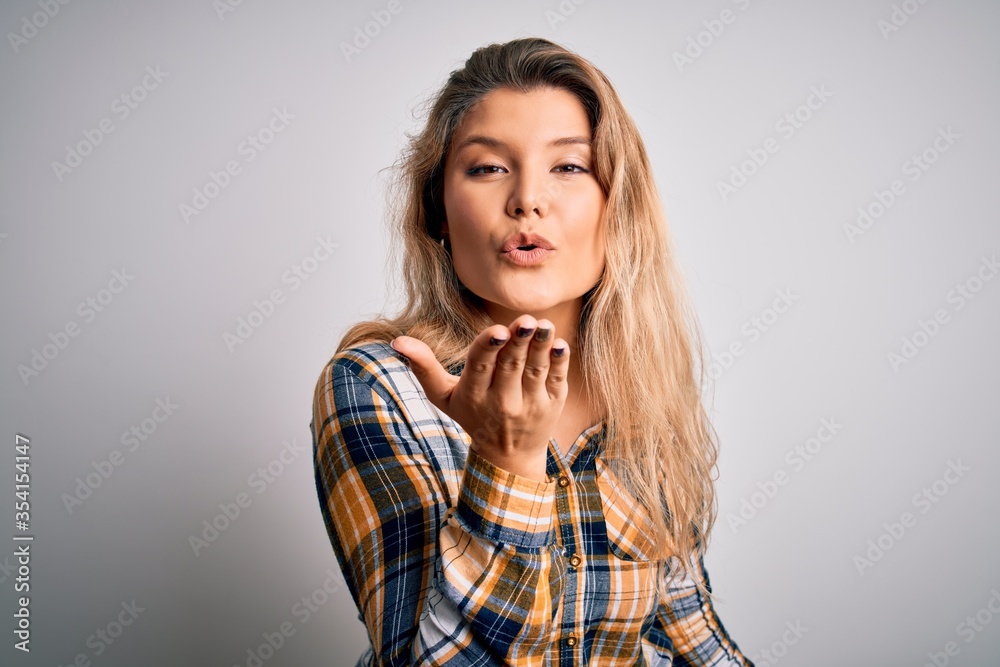 Young beautiful blonde woman wearing casual shirt standing over isolated white background looking at the camera blowing a kiss with hand on air being lovely and sexy. Love expression. Young beautiful blonde woman wearing casual shirt standing over isolated white background looking at the camera blowing a kiss with hand on air being lovely and sexy. Love expression.