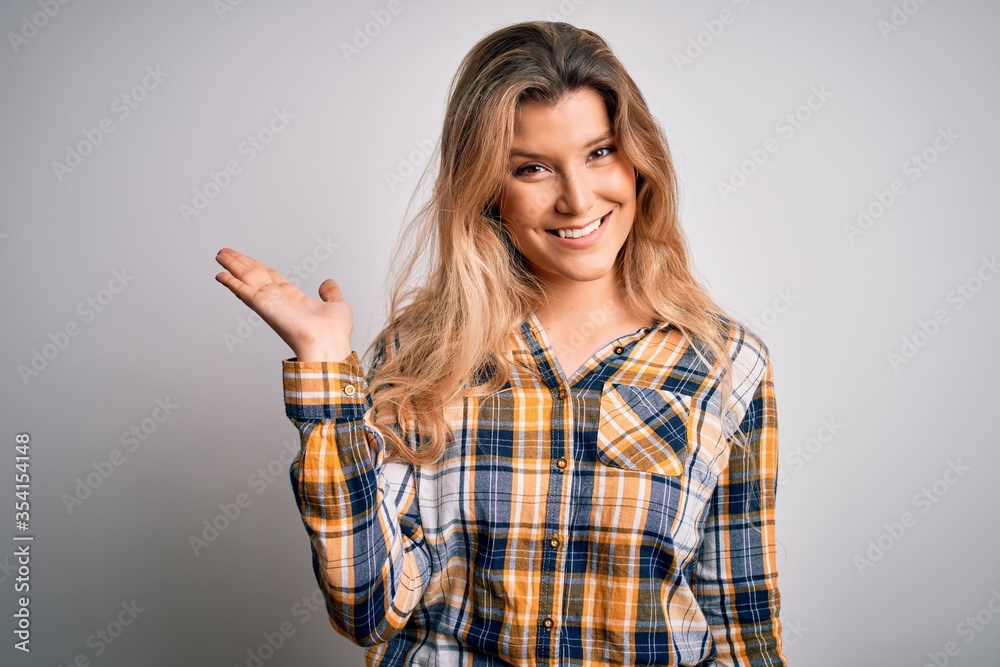 Young beautiful blonde woman wearing casual shirt standing over isolated white background smiling cheerful presenting and pointing with palm of hand looking at the camera. Young beautiful blonde woman wearing casual shirt standing over isolated white background smiling cheerful presenting and pointing with palm of hand looking at the camera.