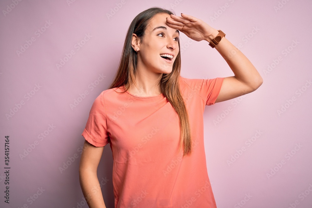 Young beautiful woman wearing casual t-shirt standing over isolated pink background very happy and smiling looking far away with hand over head. Searching concept. Young beautiful woman wearing casual t-shirt standing over isolated pink background very happy and smiling looking far away with hand over head. Searching concept.