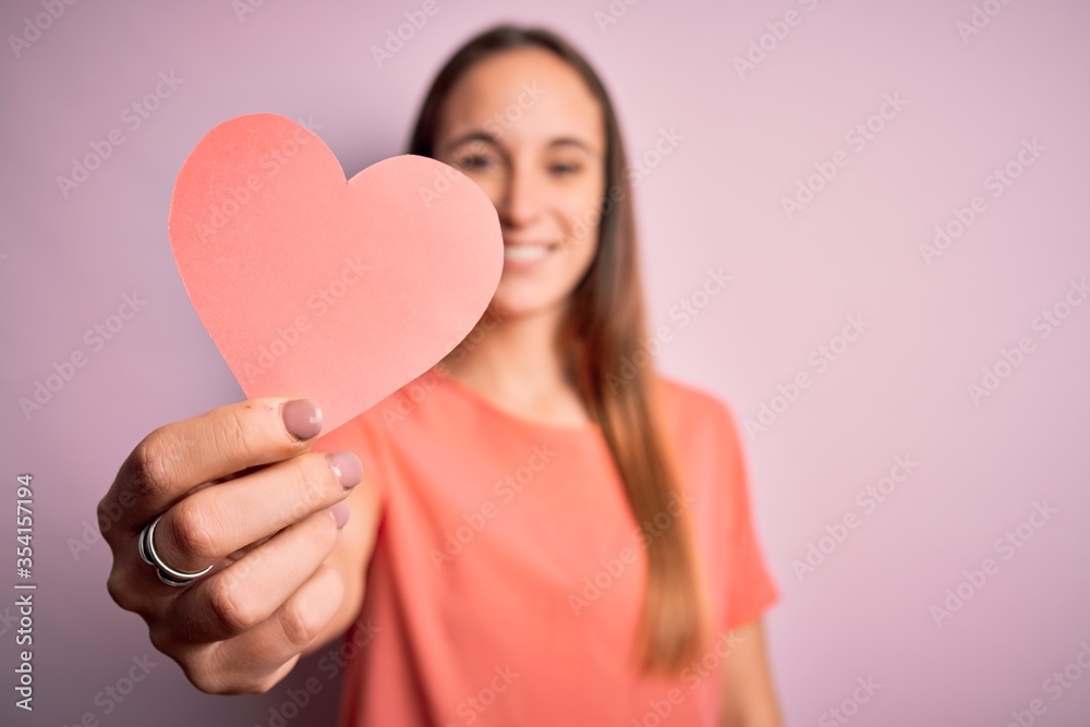 Young beautiful romantic woman holding paper heart shape over isolated pink background with a happy face standing and smiling with a confident smile showing teeth Young beautiful romantic woman holding paper heart shape over isolated pink background with a happy face standing and smiling with a confident smile showing teeth