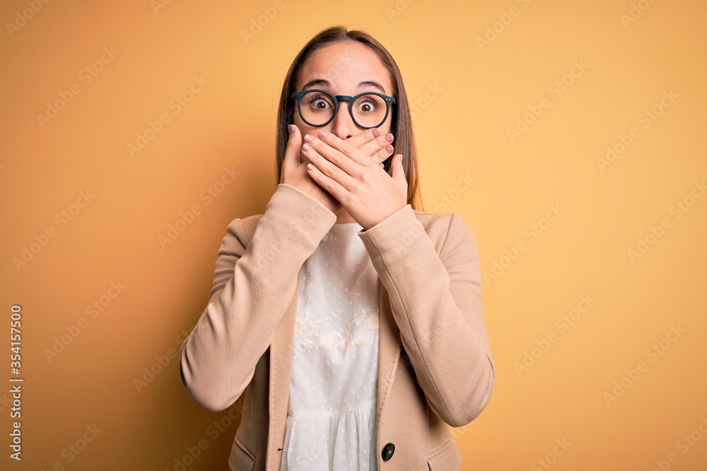 Young beautiful businesswoman wearing jacket and glasses over isolated yellow background shocked covering mouth with hands for mistake. Secret concept.