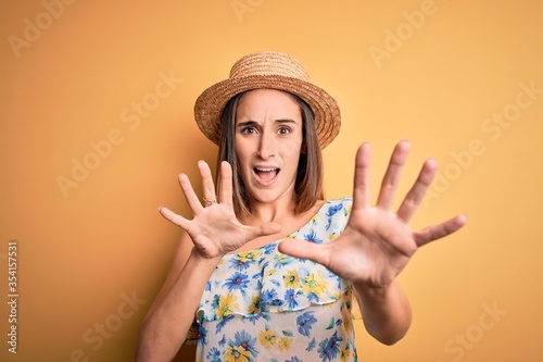 Young beautiful woman wearing casual t-shirt and summer hat over isolated yellow background afraid and terrified with fear expression stop gesture with hands, shouting in shock. Panic concept.