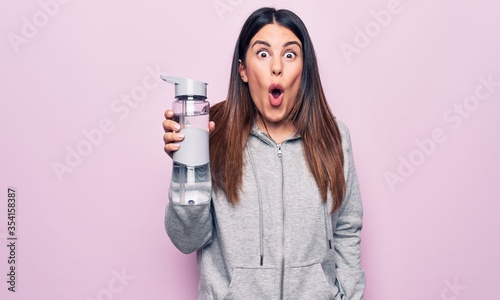 Young beautiful brunette sporty woman drinking bottle of water over isolated pink background scared and amazed with open mouth for surprise, disbelief face