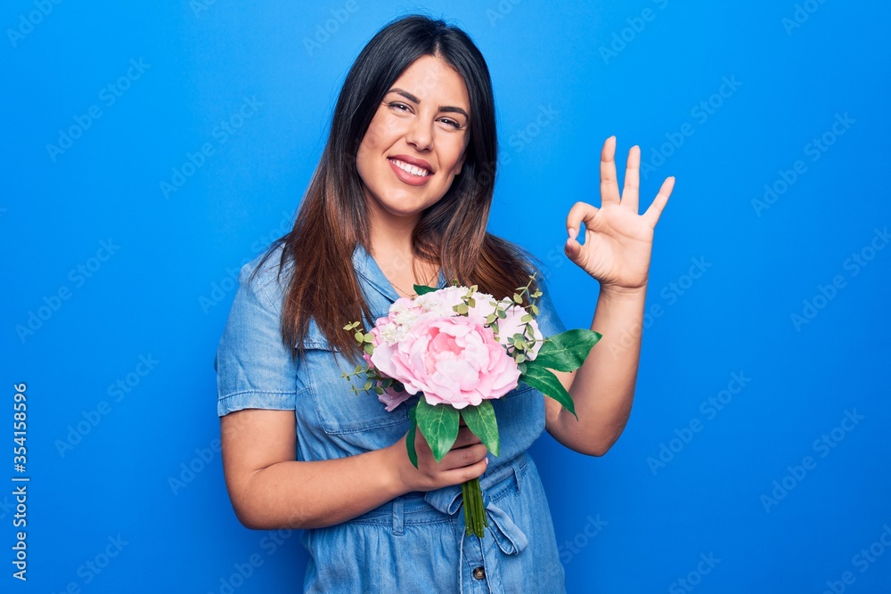 Young beautiful brunette woman holding bouquet of pink flowers over isolated blue background doing ok sign with fingers, smiling friendly gesturing excellent symbol Young beautiful brunette woman holding bouquet of pink flowers over isolated blue background doing ok sign with fingers, smiling friendly gesturing excellent symbol
