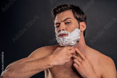Young handsome man with shaving cream on his face, grooming his beard with straight razor and looking at camera