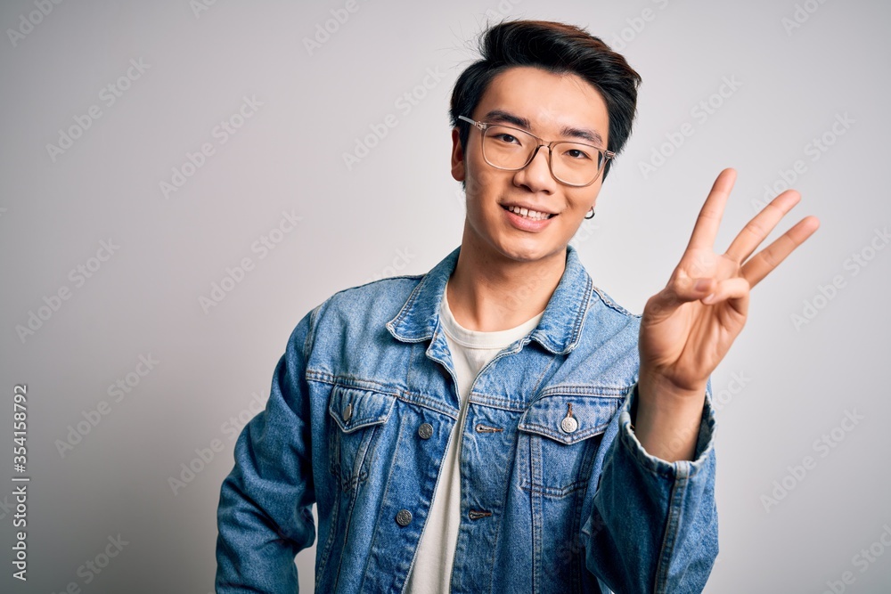 Young handsome chinese man wearing denim jacket and glasses over white background showing and pointing up with fingers number three while smiling confident and happy. Young handsome chinese man wearing denim jacket and glasses over white background showing and pointing up with fingers number three while smiling confident and happy.