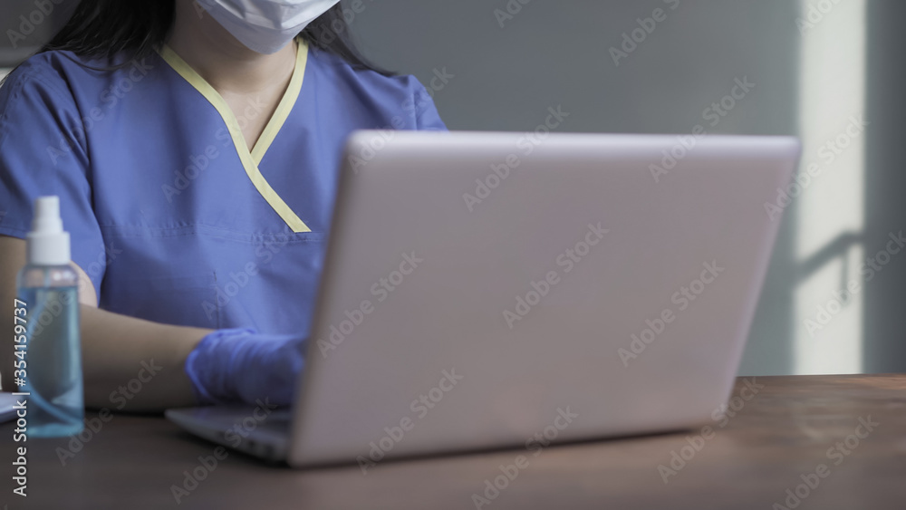 Medic working laptop sitting at table with computer and antiseptic spray on it in blurred foreground. Highlighted focus on female torso in blue uniform. Close up shot. Copy space at right side. Medic working laptop sitting at table with computer and antiseptic spray on it in blurred foreground. Highlighted focus on female torso in blue uniform. Close up shot. Copy space at right side.