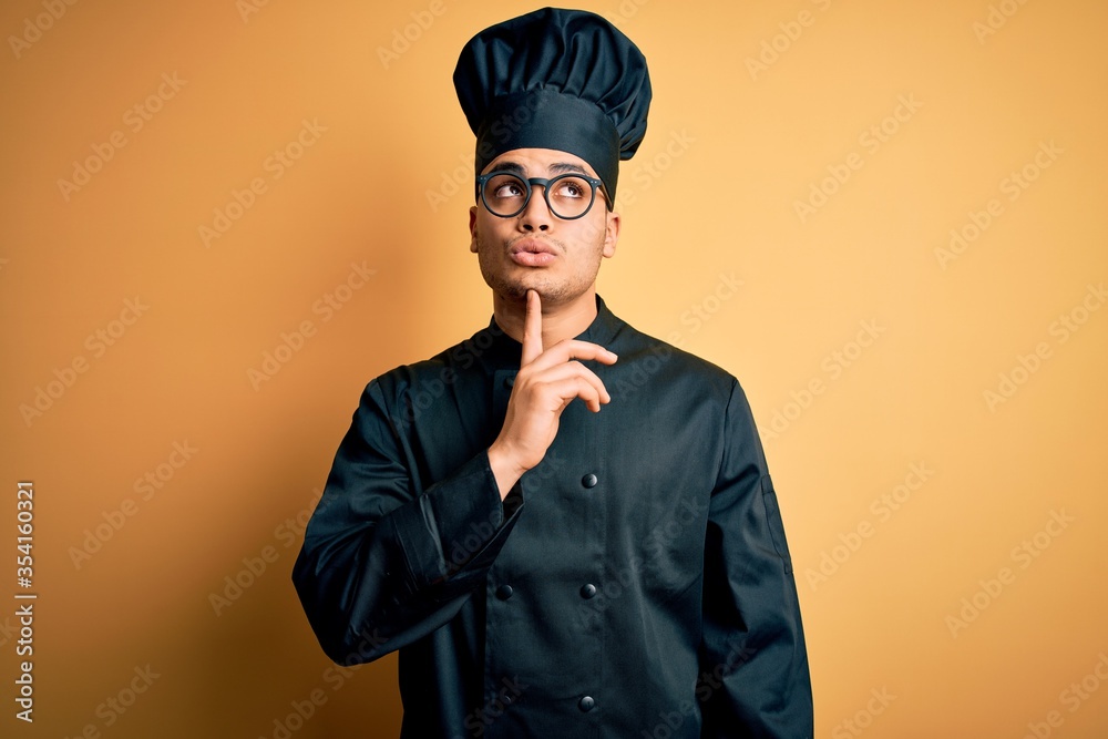 Young brazilian chef man wearing cooker uniform and hat over isolated yellow background Thinking concentrated about doubt with finger on chin and looking up wondering Young brazilian chef man wearing cooker uniform and hat over isolated yellow background Thinking concentrated about doubt with finger on chin and looking up wondering