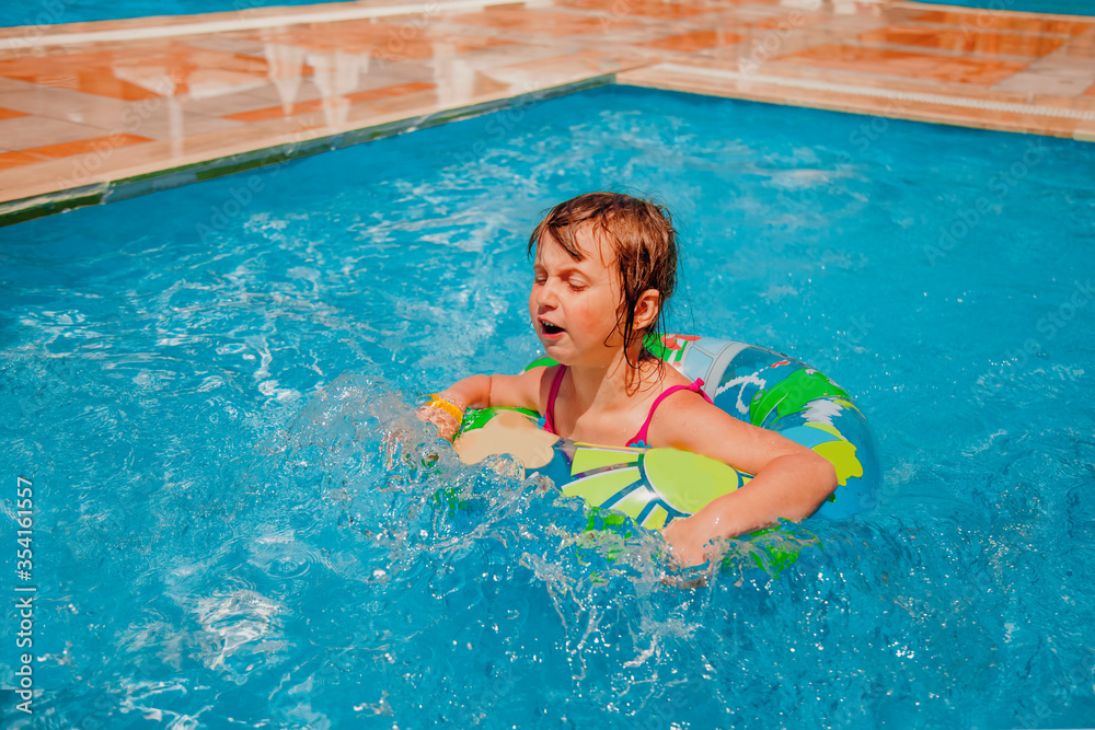 Funny facial expression of little cute child girl swimming with colorful ring outdoors in pool. Summer holiday and happy childhood concept.