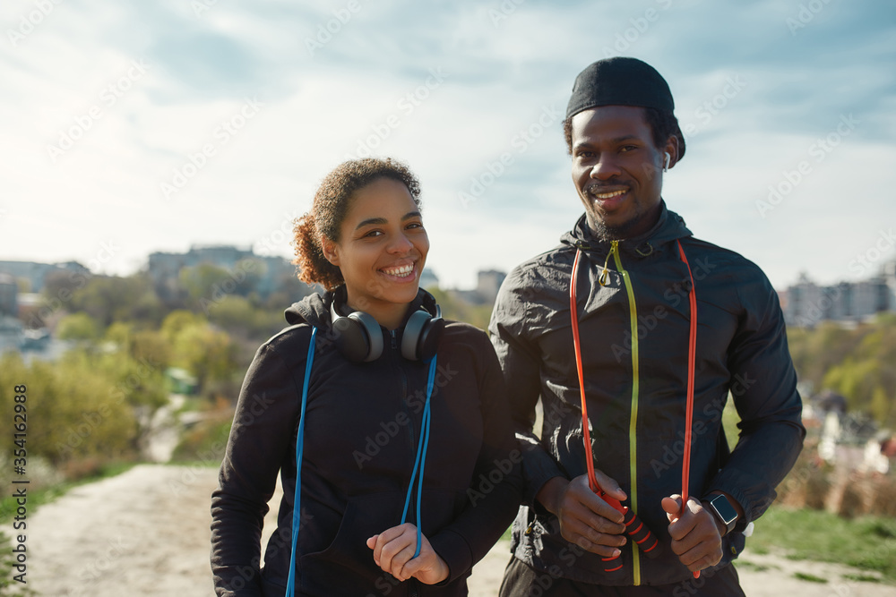 Beautiful young african couple in sportswear holding jumping ropes, looking at camera and smiling while working out together outdoors