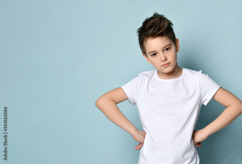 Brunet teenage boy in white t-shirt. He put his hands on hips and looking at you, posing against blue studio background