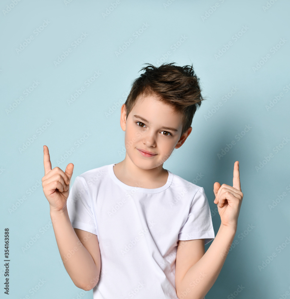 Nice amiable little boy in white t-shirt looking forward with smile putting both index fingers up. Half-length portrait isolated on light blue Nice amiable little boy in white t-shirt looking forward with smile putting both index fingers up. Half-length portrait isolated on light blue
