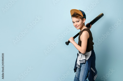 Sporty little boy in sleeveless shirt, jeans and cap preparing to hit with baseball bat from his shoulder. Side view shot isolated on blue background