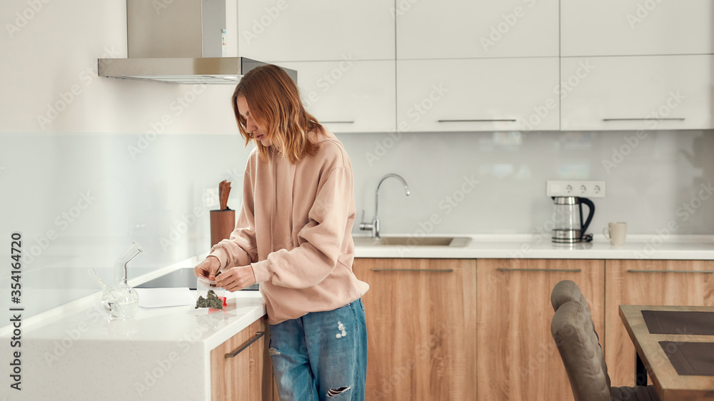 Impove your day. Woman getting cannabis buds out of a plastic bag for grinding them using red marijuana grinder, while standing in the kitchen. Glass water pipe or bong on the table Impove your day. Woman getting cannabis buds out of a plastic bag for grinding them using red marijuana grinder, while standing in the kitchen. Glass water pipe or bong on the table