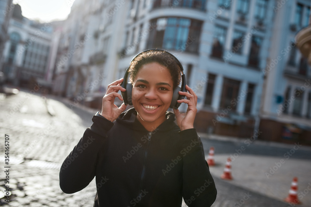 Best jogging music. Happy african female runner wearing headphones looking at camera and smiling. Young woman in sportswear running outdoors. Morning jog