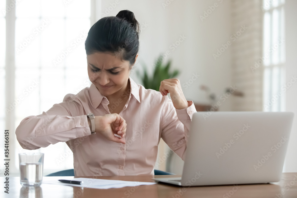 Stressed young indian businesswoman sitting in office, looking in hurry at wristwatch, irritated by non-punctual video call from clients. Unhappy millennial woman checking time to workday finish. Stressed young indian businesswoman sitting in office, looking in hurry at wristwatch, irritated by non-punctual video call from clients. Unhappy millennial woman checking time to workday finish.
