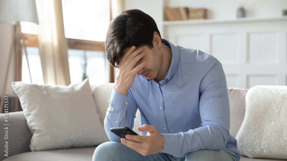 Upset young man sit on sofa at home frustrated by negative message on smartphone gadget, unhappy male distressed by eviction notice email or bad news on cellphone, get cell problems or virus Upset young man sit on sofa at home frustrated by negative message on smartphone gadget, unhappy male distressed by eviction notice email or bad news on cellphone, get cell problems or virus
