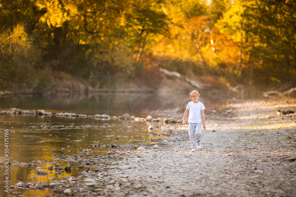 Little blonde boy in green glasses playing on the riverside. Autumn in the yellow forest Little blonde boy in green glasses playing on the riverside. Autumn in the yellow forest