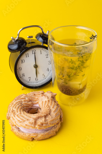 A glass of buckwheat tea with choux ring bun on minimalist bright yellow background, with black analog clock