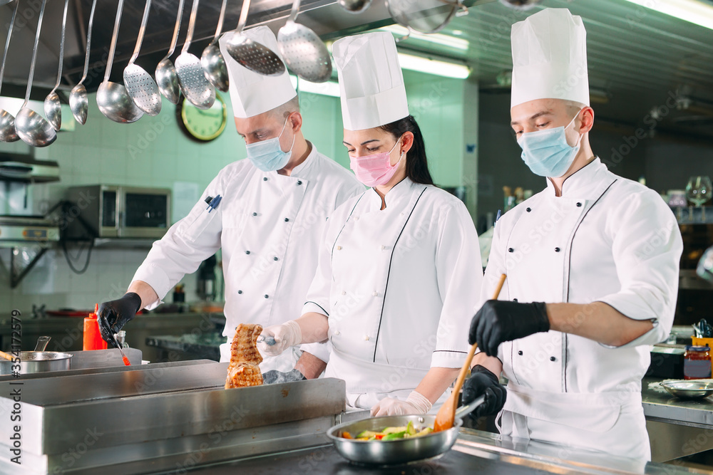 Chefs in protective masks and gloves prepare food in the kitchen of a restaurant or hotel. Chefs in protective masks and gloves prepare food in the kitchen of a restaurant or hotel.