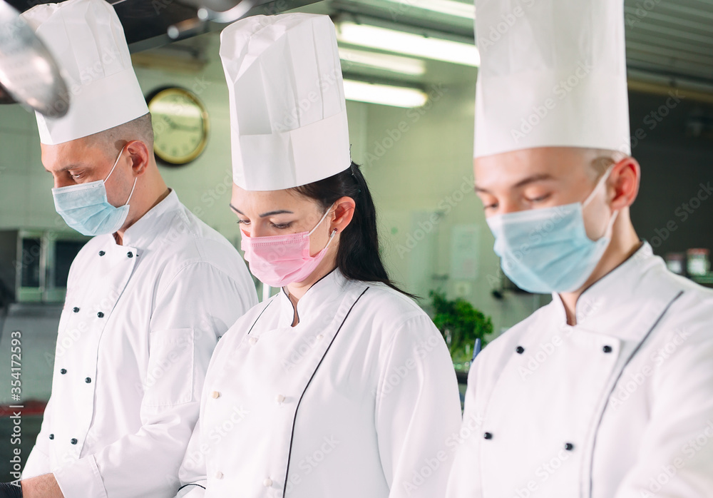 Chefs in protective masks and gloves prepare food in the kitchen of a restaurant or hotel.