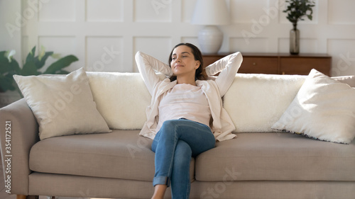 Happy young Caucasian woman sit relax on comfortable sofa at home look in window distance thinking dreaming, smiling millennial girl rest on couch, breathe fresh air, stress free, relaxation concept