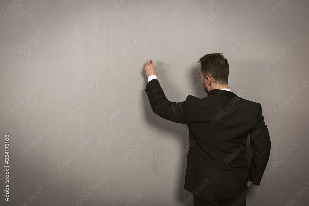 Rear view of businessman writing on wall. Creative man in black elegance suit drawing by pan or chalk on light grey wall. Template concept. Toned image. Rear view of businessman writing on wall. Creative man in black elegance suit drawing by pan or chalk on light grey wall. Template concept. Toned image.