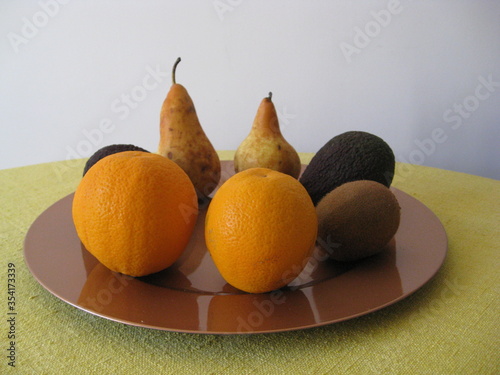 varias frutas: dos peras, dos aguacates, dos kivis, dos naranjas cítricos, carnosas y de agua, ecológicas en plato rosa, sobre mesa y fondo blanco pared. Vista frontal
 photo