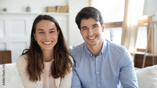Headshot portrait of smiling young caucasian man and woman sit on sofa in living room having web video call, happy millennial couple have fun talk speak on webcam internet conference at home together