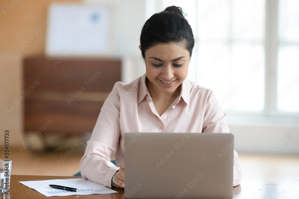 Head shot positive indian businesswoman working on laptop, sitting at desk in modern office. Smiling female hindu financial analyst writing economic research, consulting clients online at workplace. Head shot positive indian businesswoman working on laptop, sitting at desk in modern office. Smiling female hindu financial analyst writing economic research, consulting clients online at workplace.