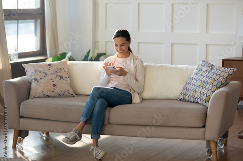 Smiling millennial girl relax on cozy couch at home texting messaging on modern cellphone gadget, happy young Caucasian woman sit rest on sofa in living room browse wireless internet shopping on cell