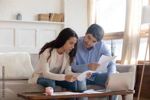 Focused millennial caucasian couple sit on sofa at home manage household paperwork finances document together, young family pay bills taxes on laptop online, consider paper correspondence