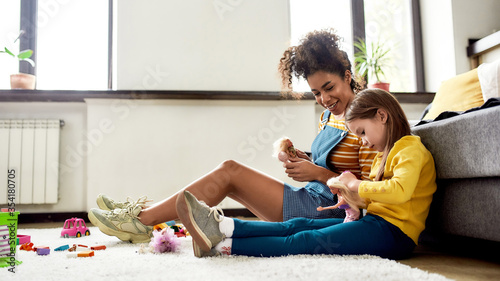 Kidspace. African american woman baby sitter entertaining caucasian cute little girl. They are playing with dolls, sitting on the floor photo