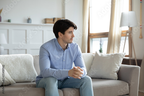 Thoughtful young caucasian man sit on sofa in living room look in window distance pondering or planning, pensive millennial male lost in thoughts thinking or dreaming at home, rest on couch on weekend