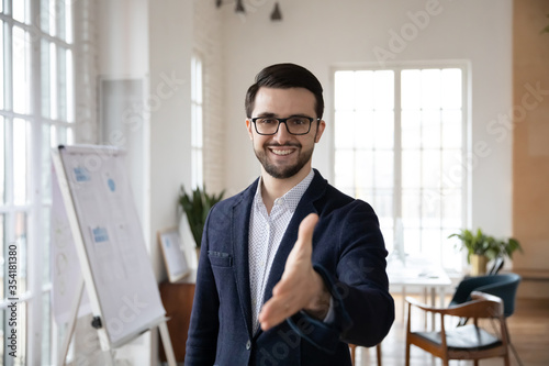 Head shot pleasant smiling trustful manager in suit reaching out hand, looking at camera. Happy confident young businessman welcoming greeting clients partners in modern office, acquaintance concept.