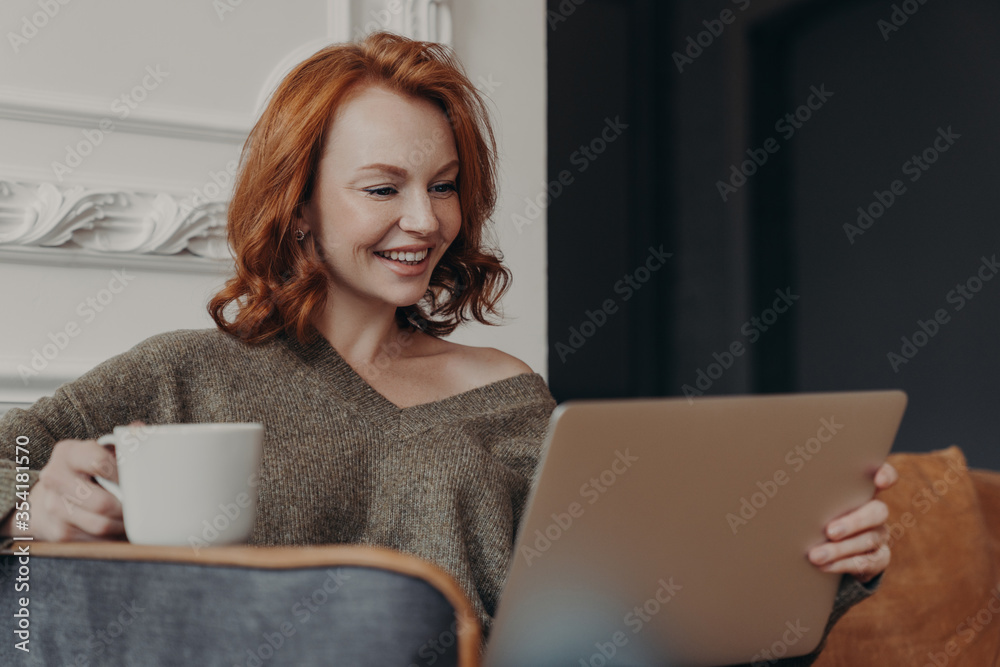 Beautiful positive redhead woman happy to have video call with best friend, share news with each other, drinks coffee, sits in comfortable armchair, watches intereting movie, connected to internet Beautiful positive redhead woman happy to have video call with best friend, share news with each other, drinks coffee, sits in comfortable armchair, watches intereting movie, connected to internet