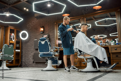 Man visiting barbershop. Side view of professional barber is drying hair of his young client who is sitting in chair in front of a mirror in the barbershop