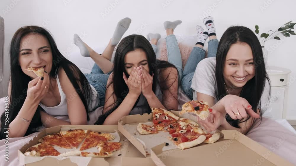 Three gorgeous girlfriends are lying on the bed. The women are eating takeaway pizza and talking. They are having fun together. Three gorgeous girlfriends are lying on the bed. The women are eating takeaway pizza and talking. They are having fun together.