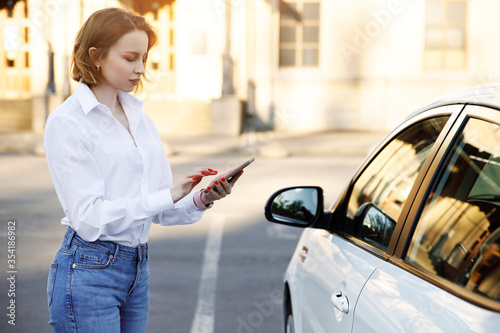 Woman using mobile phone, communication or online application, standing near car on city street or parking, outdoors. Car sharing, rental service or taxi app.