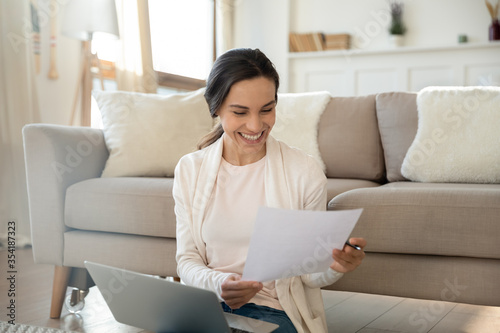 Smiling young Caucasian woman sit in living room feel excited reading good news in paper letter, happy millennial girl work on laptop at home, laugh get pleasant message in paperwork correspondence