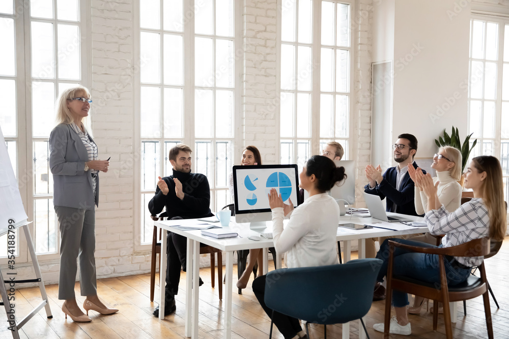 Full length satisfied with good presentation young diverse businesspeople applauding to middle aged pleasant smart leader speaker coach trainer mentor standing near flipchart in modern office. Full length satisfied with good presentation young diverse businesspeople applauding to middle aged pleasant smart leader speaker coach trainer mentor standing near flipchart in modern office.