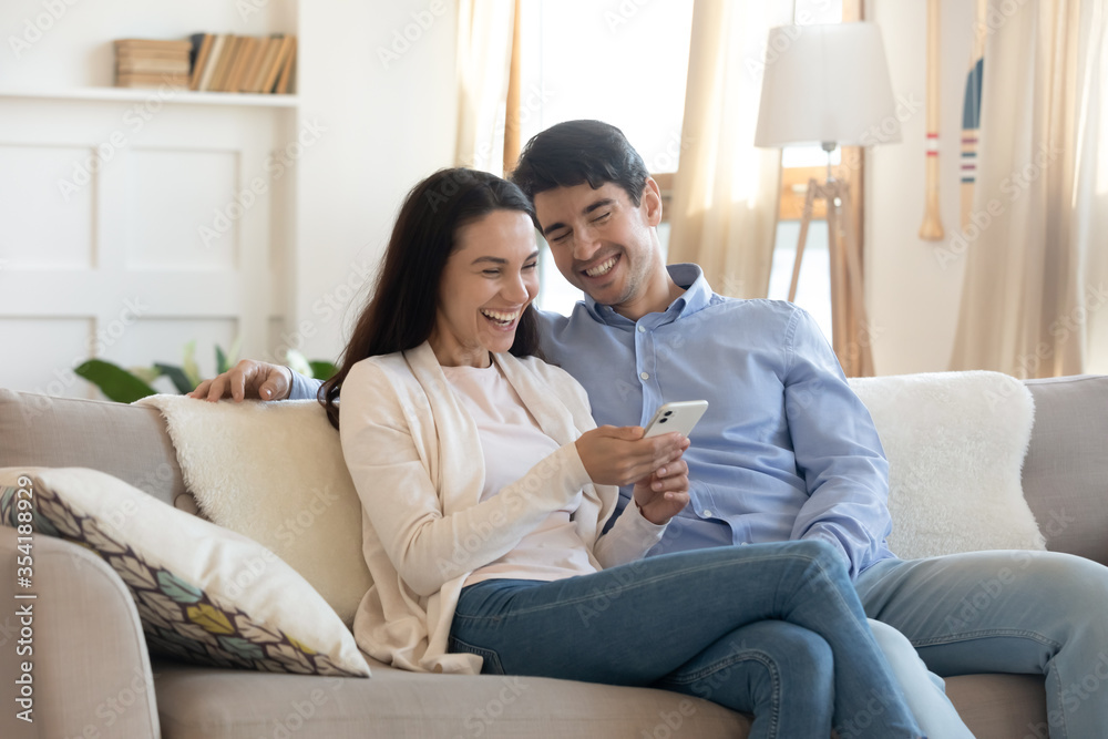 Overjoyed young caucasian man and woman sit rest on couch in living room have fun using smartphone together, happy millennial couple laugh watching funny video on cellphone, enjoy weekend indoors Overjoyed young caucasian man and woman sit rest on couch in living room have fun using smartphone together, happy millennial couple laugh watching funny video on cellphone, enjoy weekend indoors