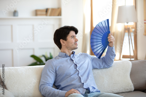 Overheated young caucasian man sit rest on sofa in living room breathe fresh air using hand waver, exhausted millennial male feel heatstroke suffer from hot weather, use fan relaxing on couch