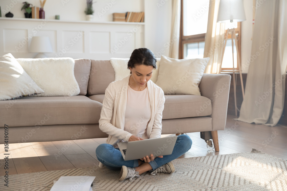 Concentrated millennial Caucasian girl sit on floor in living room working or studying on laptop, focused young woman busy preparing report or paying bills online on computer, education concept Concentrated millennial Caucasian girl sit on floor in living room working or studying on laptop, focused young woman busy preparing report or paying bills online on computer, education concept