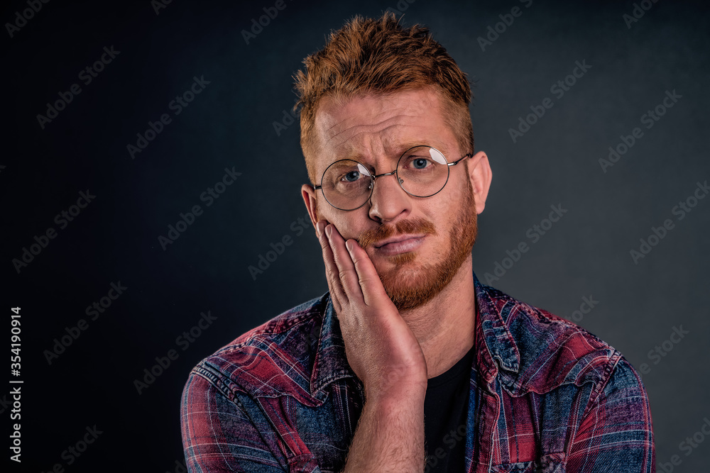 Headshot of upset and gloomy redhead guy with beard in transparent glasses, frowning holding palm on cheek gazing with regret and disappointment, losing chance over gray background Headshot of upset and gloomy redhead guy with beard in transparent glasses, frowning holding palm on cheek gazing with regret and disappointment, losing chance over gray background
