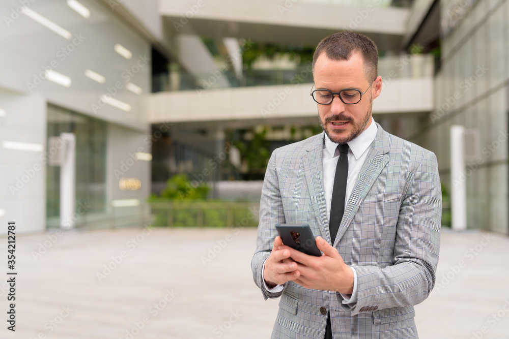 Hispanic bald bearded businessman using phone with eyeglasses in the city outdoors Hispanic bald bearded businessman using phone with eyeglasses in the city outdoors