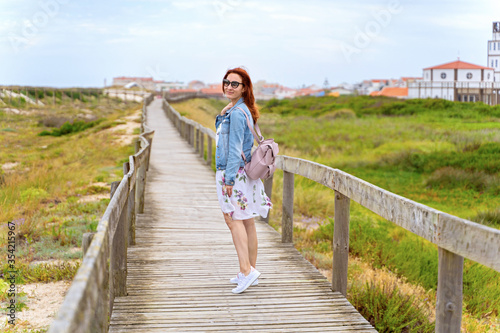 Red head woman posing in white dress and jean jacket on beach wooden board walk. Smiling tourist enjoying summer vacation in Costa Nova in Aveiro, Portugal. Travel destination concept © Elena Sistaliuk